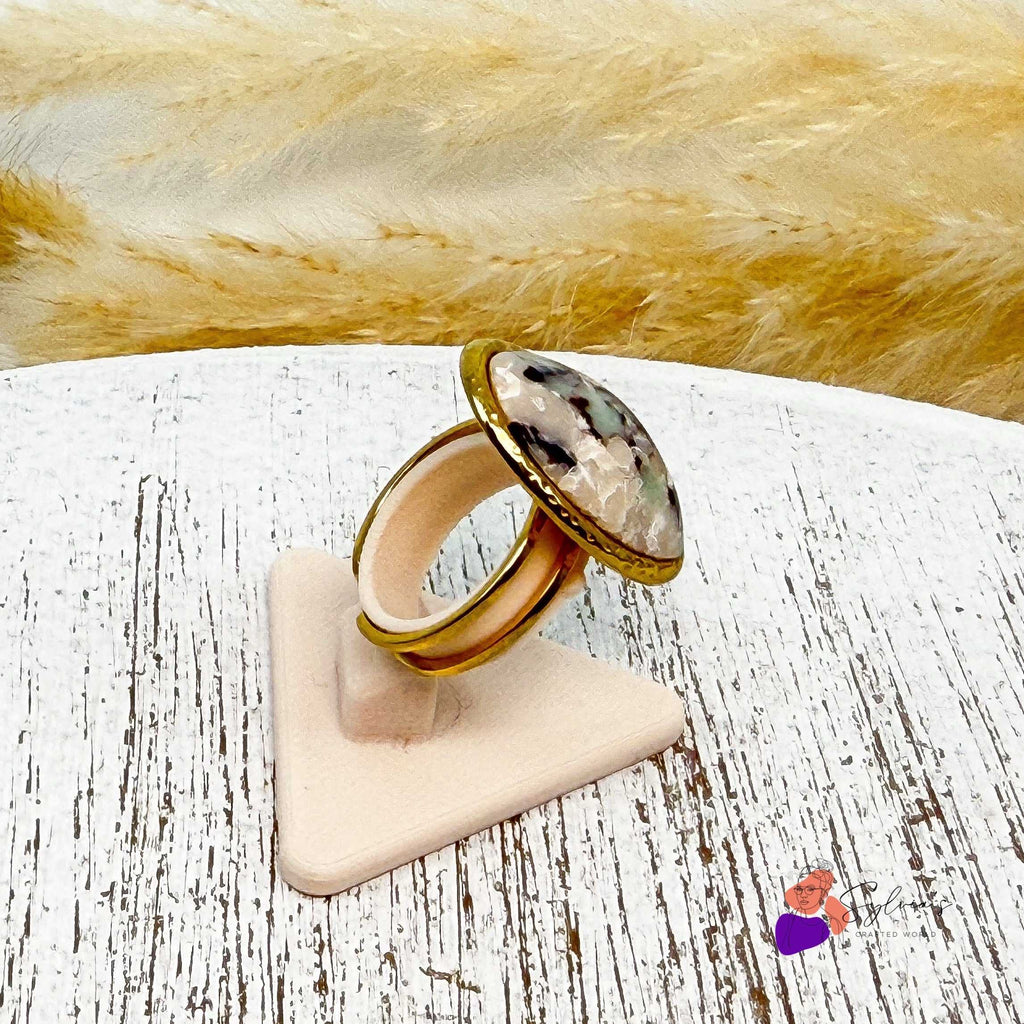 Bague artisanale ovale Clara en argile polymère marbrée et acier doré, montée sur support ajustable, exposée sur présentoir beige texturé.