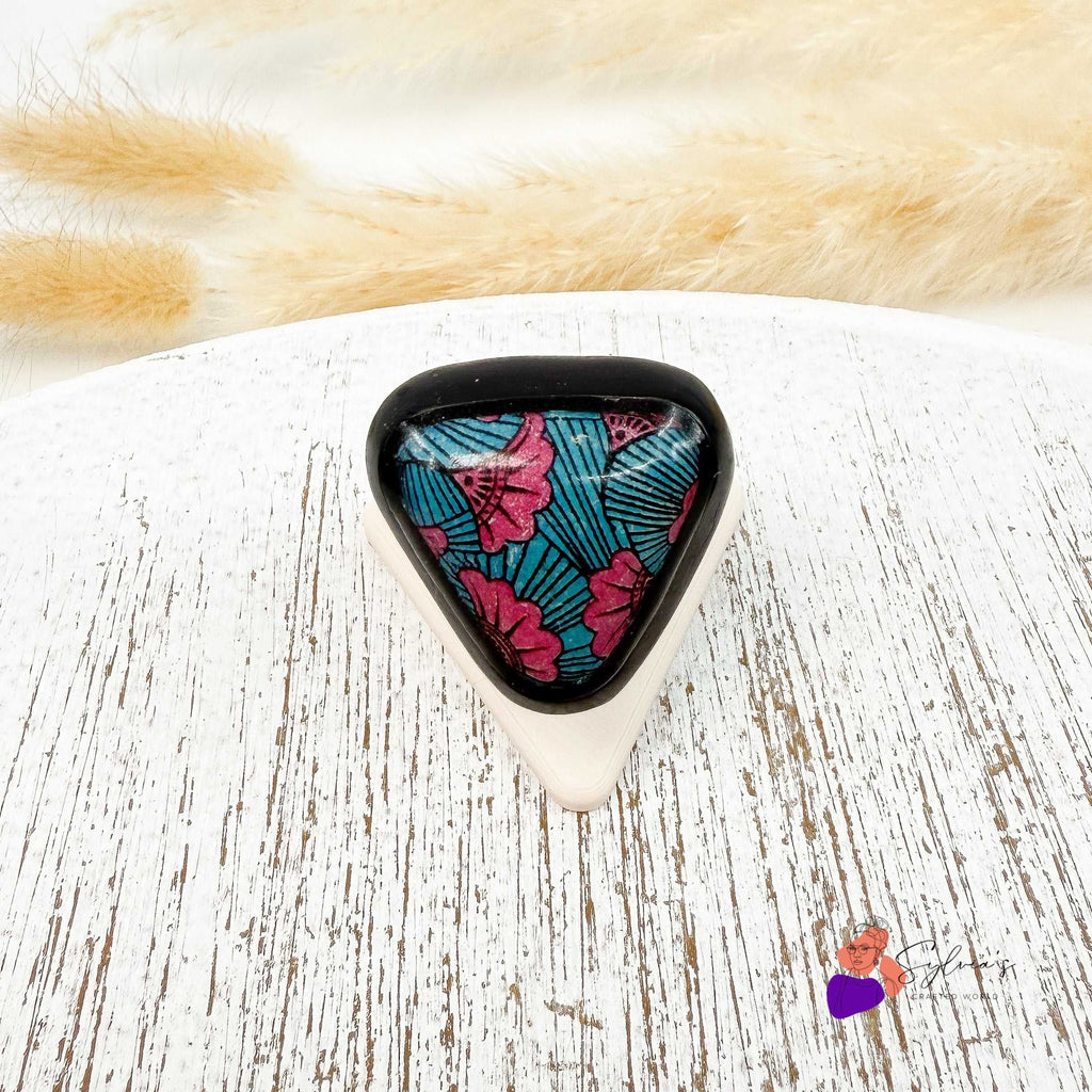 Bague artisanale motif wax en argile polymère 'Aya' aux couleurs vives, forme triangulaire avec fleurs roses et fond bleu sur support blanc.