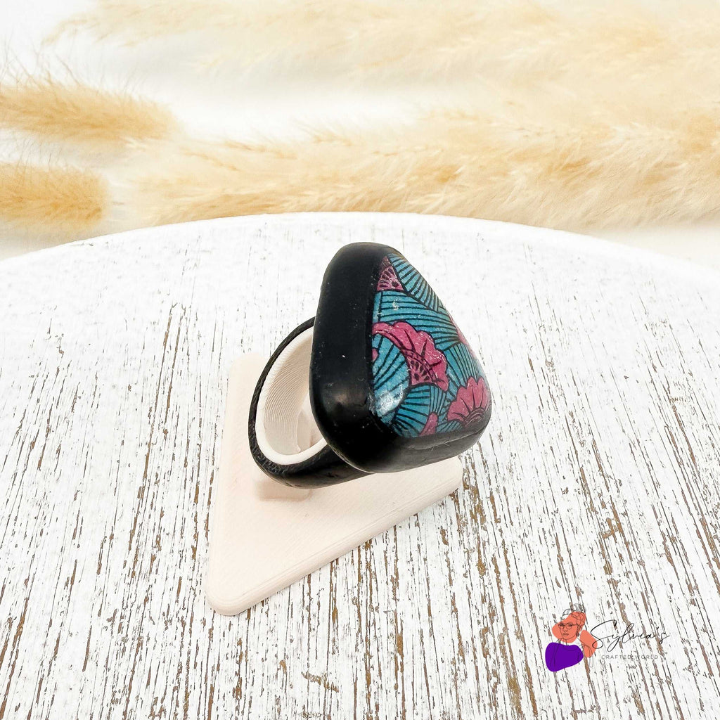 Bague artisanale motif wax en argile polymère 'Aya' aux couleurs vives, forme triangulaire sur support noir, bijou original et coloré.