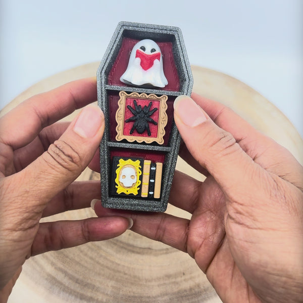 Vidéo du porte-clé Cercueil spooky imprimé en 3D, en forme de cercueil noir pailleté avec fantôme blanc, araignée encadrée, livres miniatures et anneau doré sur chaîne dorée. Les éléments sur les étagères sont pressés pour montrer la fonction cliquable.